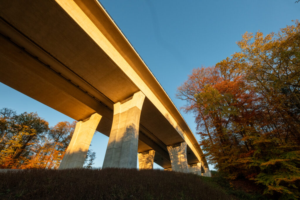 Betonbrücke mit mehreren Stützen, unter einem klaren blauen Himmel und umgeben von herbstlichen Bäumen.