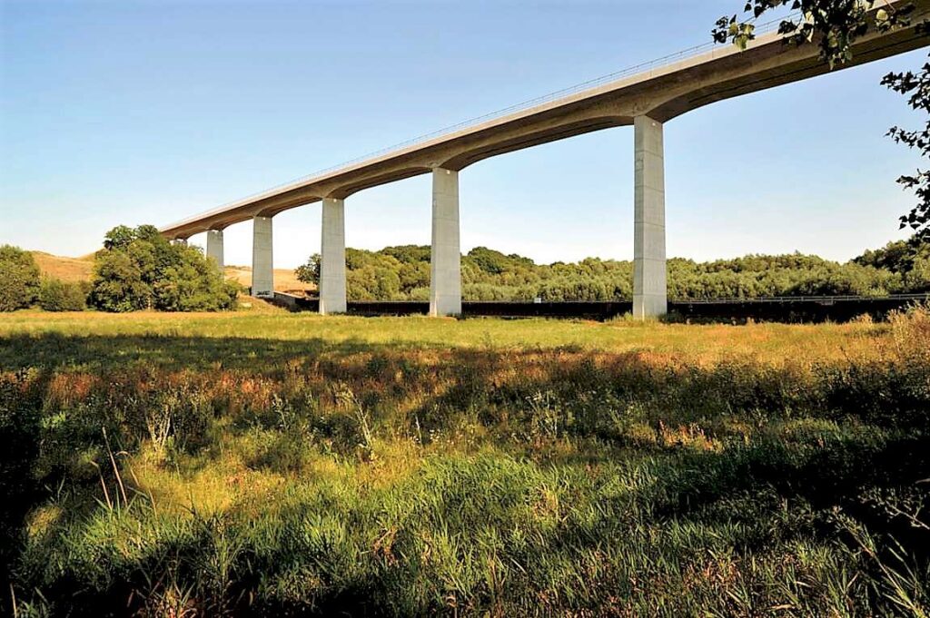 Brücke mit Betonpfeilern, die über eine Wiese führt, umgeben von Bäumen und Grünflächen.
