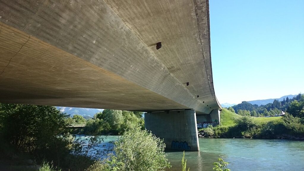 Betonbrücke mit sichtbarem Unterbau und Wasser darunter, umgeben von Vegetation.