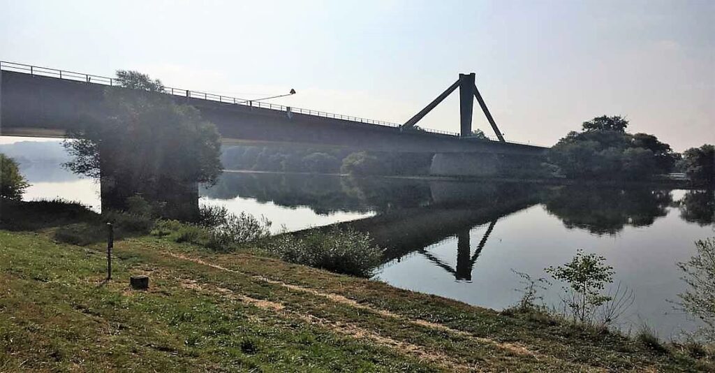 Brücke mit einem hohen Pfeiler, die sich über einen ruhigen Fluss erstreckt, mit reflektierendem Wasser im Vordergrund.