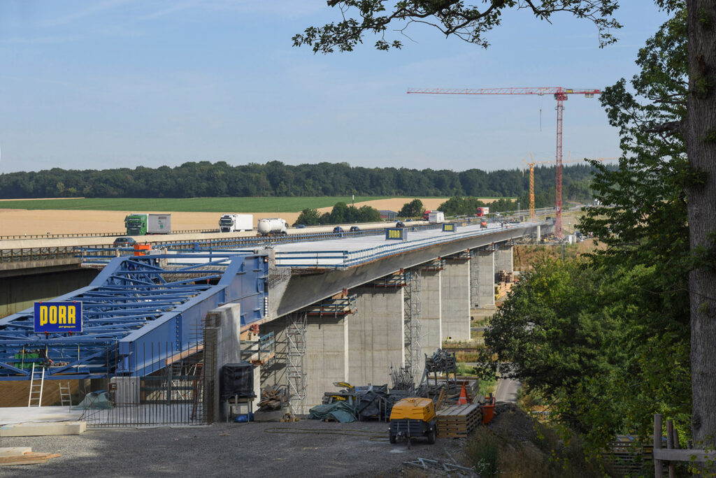 Bau einer Brücke mit blauen Stützen und Fahrzeugen auf der Fahrbahn, umgeben von Baustellenmaterial.