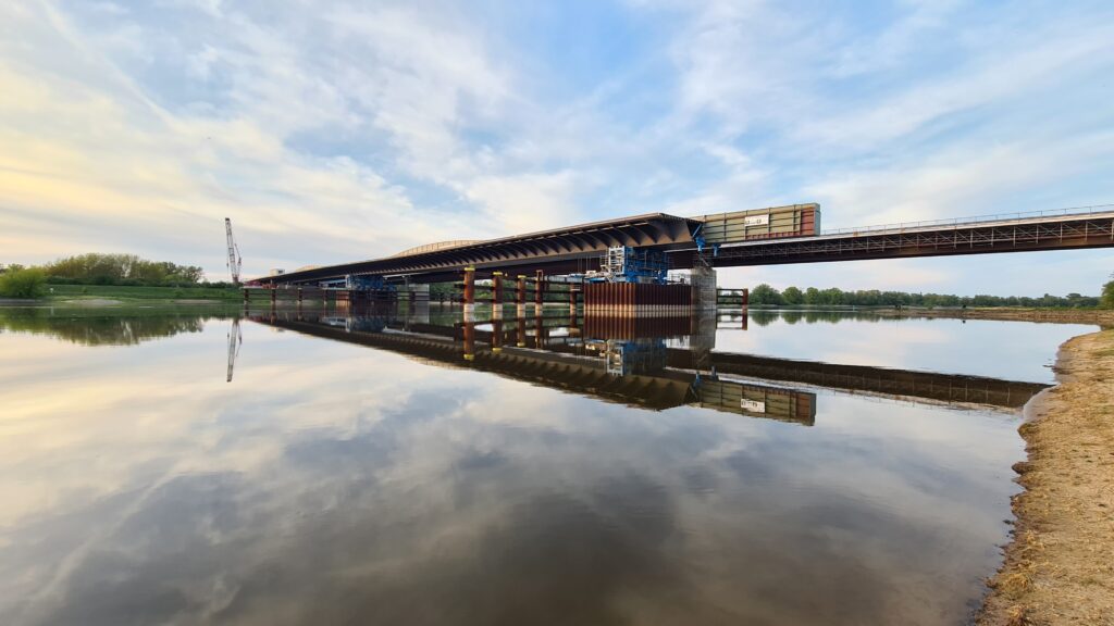 Brücke über einen ruhigen Fluss mit reflektierenden Wasseroberfläche und Bauarbeiten im Hintergrund.