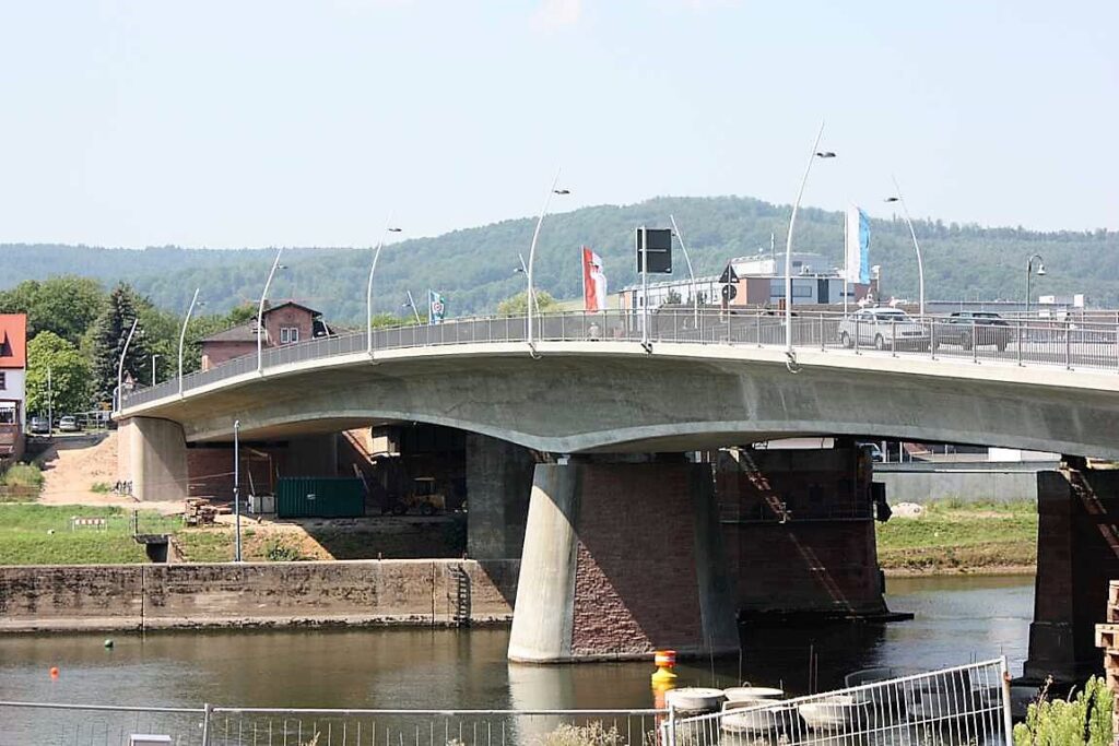 Betonbrücke mit geschwungener Form über einem Gewässer, umgeben von Grünflächen und Gebäuden.