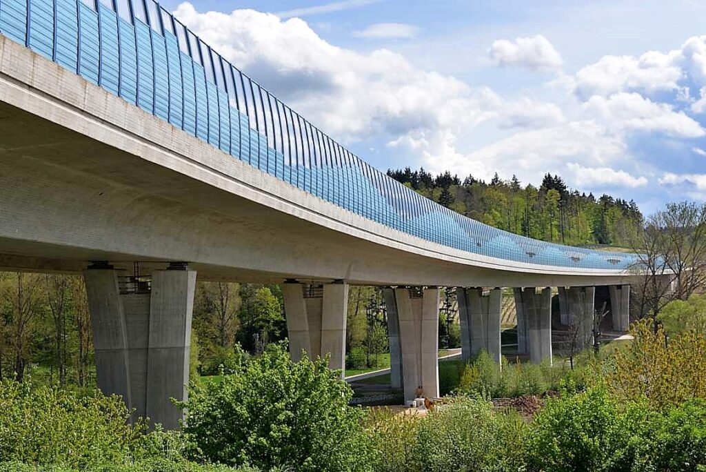 Moderne Brücke mit geschwungener Struktur und blauen Glasdetails, unterstützt von Betonsäulen.