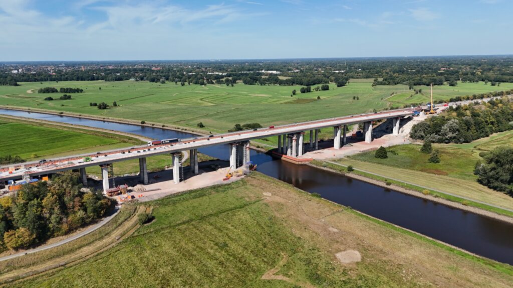 Brücke im Bau über einen Fluss mit grünen Wiesen und Bäumen in der Umgebung.