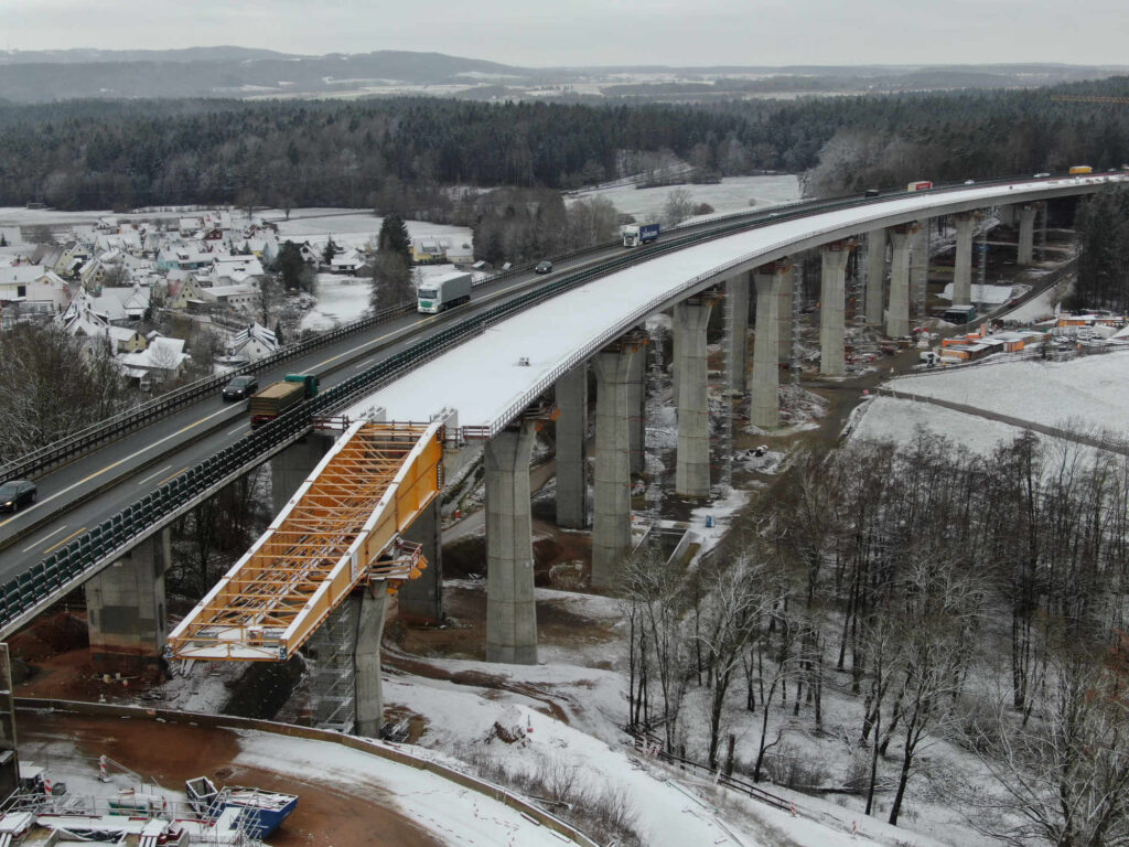 A bridge under construction with vehicles traveling on the roadway and snow on the ground.