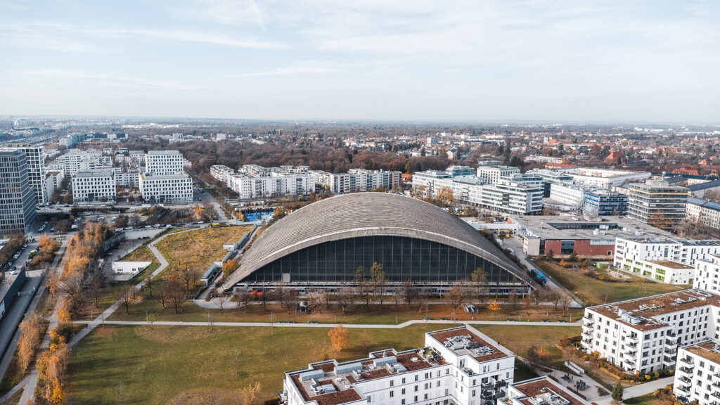 Blick auf die Paketposthalle München mit gebogenem Dach und Glasfassade, umgeben von Wohn- und Gewerbebauten im Herbst, Himmel über der Stadt